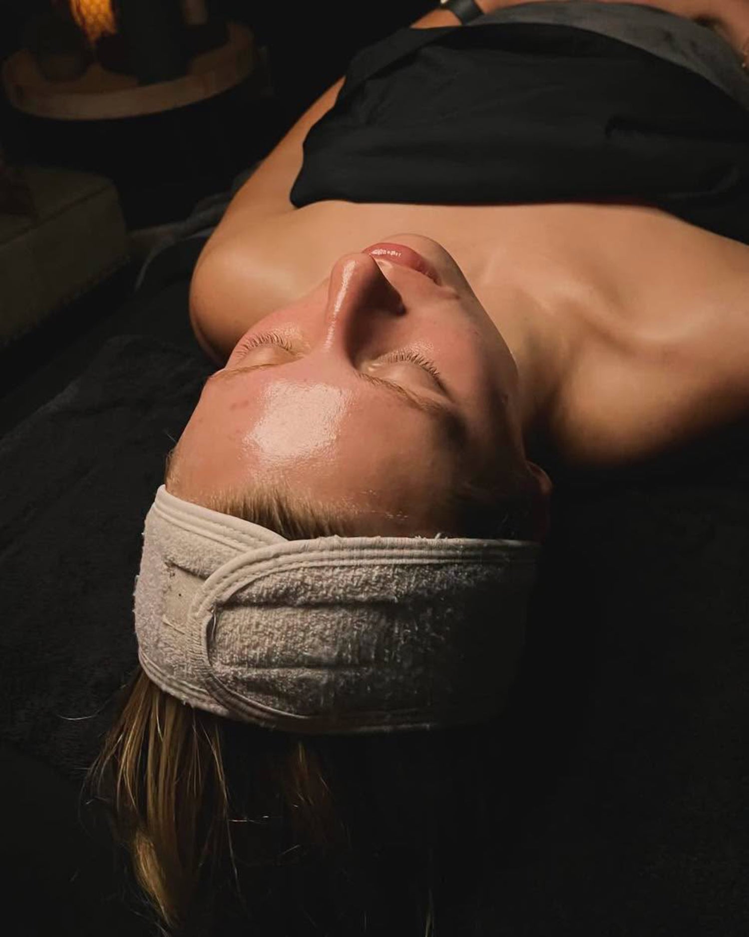 A woman lying down on a spa or massage table, wearing a light gray textured headband and a black top. Her face is wet with oil, and the background is dimly lit in black.