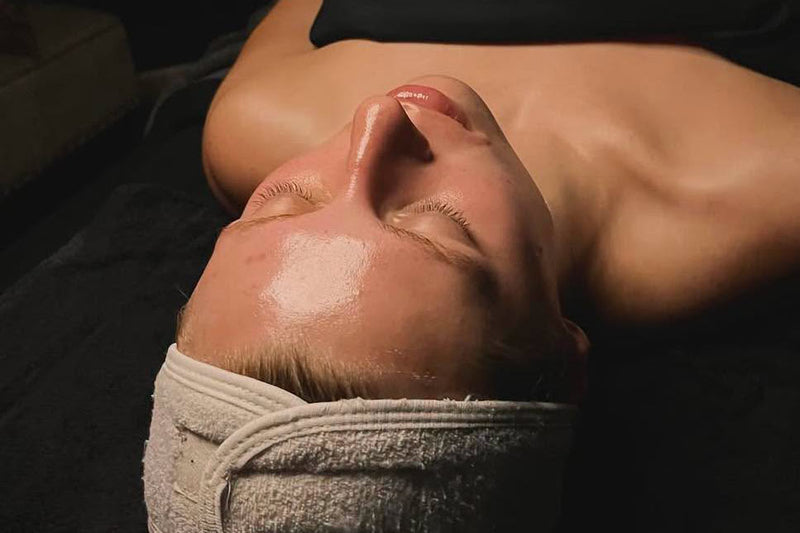 A close-up lifestyle shot of a person lying down with their eyes closed, wearing a white towel wrapped around their head, with a glossy, hydrated skin appearance under warm, dim lighting.